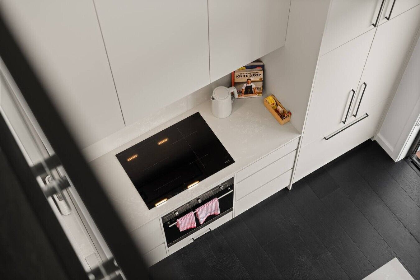 A modern kitchen with a built-in stove, a microwave, and a small oven. There are white cabinets and a countertop with a box of kitchen utensils.