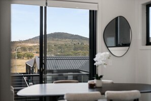 A room with a large window, a round mirror, and a table with chairs. - Cozy Alpine Retreat | Fireplace & Mountain Charm (Jindabyne)