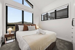 A hotel room with a large bed, a nightstand with a lamp, a window with a view of the hills, and a balcony with a railing. - Cozy Alpine Retreat | Fireplace & Mountain Charm (Jindabyne)