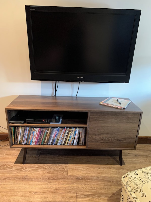 A flat-screen TV mounted on a wooden entertainment center with a shelf below containing DVDs and a remote control.