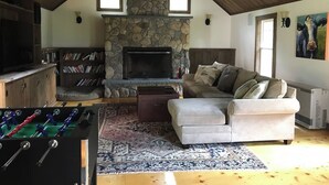 A cozy living room with a stone fireplace, a sofa, a foosball table, and a television. - Historic VT Retreat | Hot Tub, Views, Near Skiing (Mount Holly)