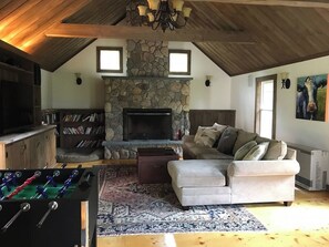 A cozy living room with a stone fireplace, a sofa, a foosball table, and a television. - Historic VT Retreat | Hot Tub, Views, Near Skiing (Mount Holly)