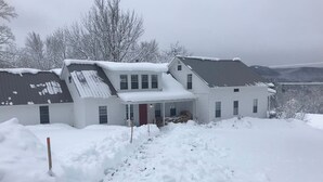 A two-story house with a snow-covered roof and a front porch. - Historic VT Retreat | Hot Tub, Views, Near Skiing (Mount Holly)