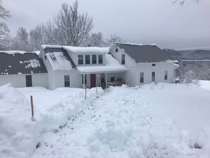A two-story house with a snow-covered roof and a front porch. - Historic VT Retreat | Hot Tub, Views, Near Skiing (Mount Holly)