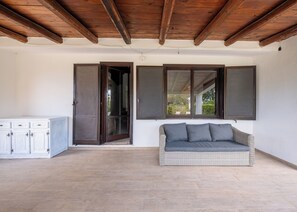 Ein Zimmer mit Holzbalkendecke, einem weißen Schrank, einem Korbsessel mit Kissen und zwei Fenstern mit Fensterläden.