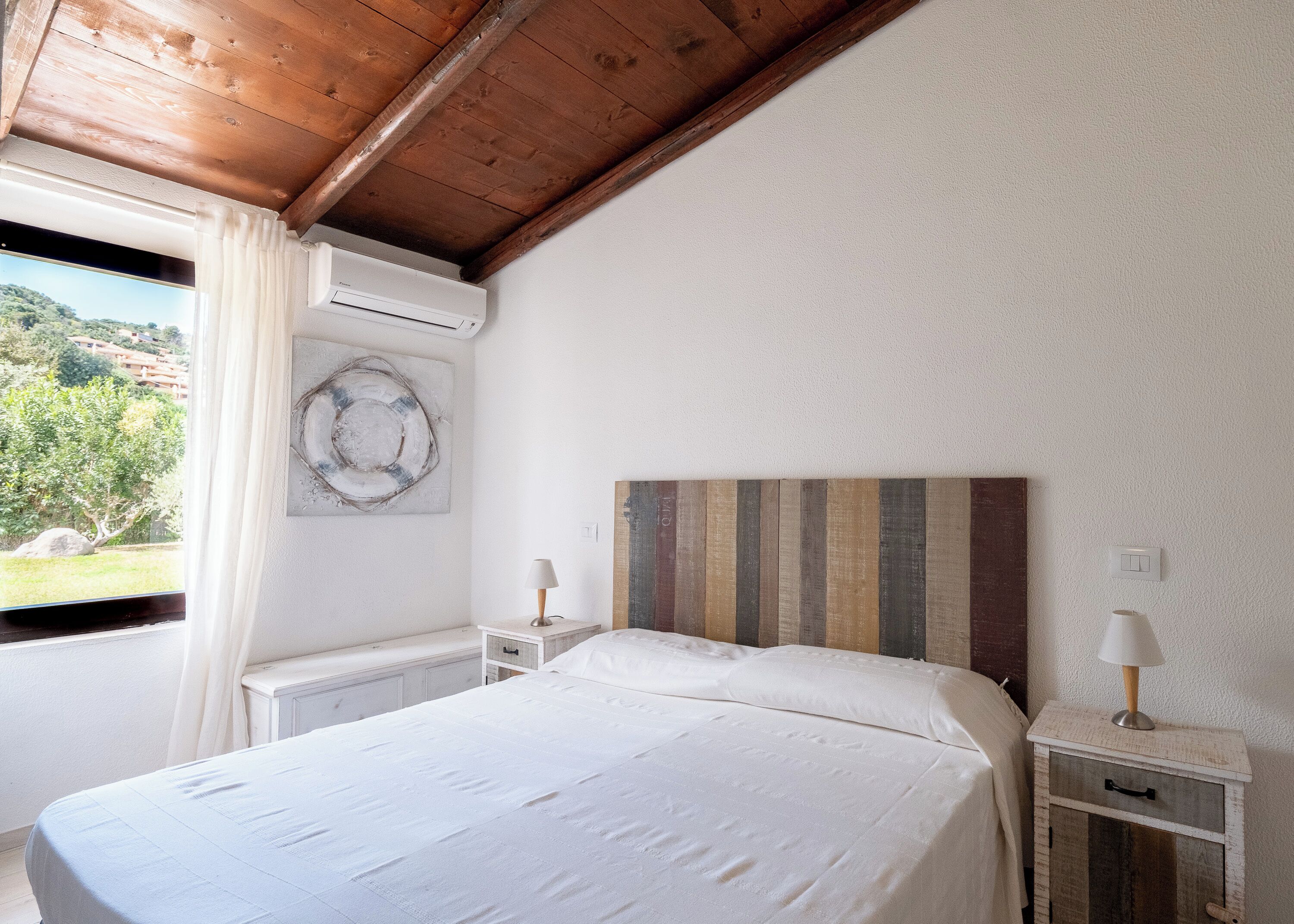 A bedroom with a wooden ceiling, a bed with white bedding, a nightstand with a lamp, and a view of greenery outside.