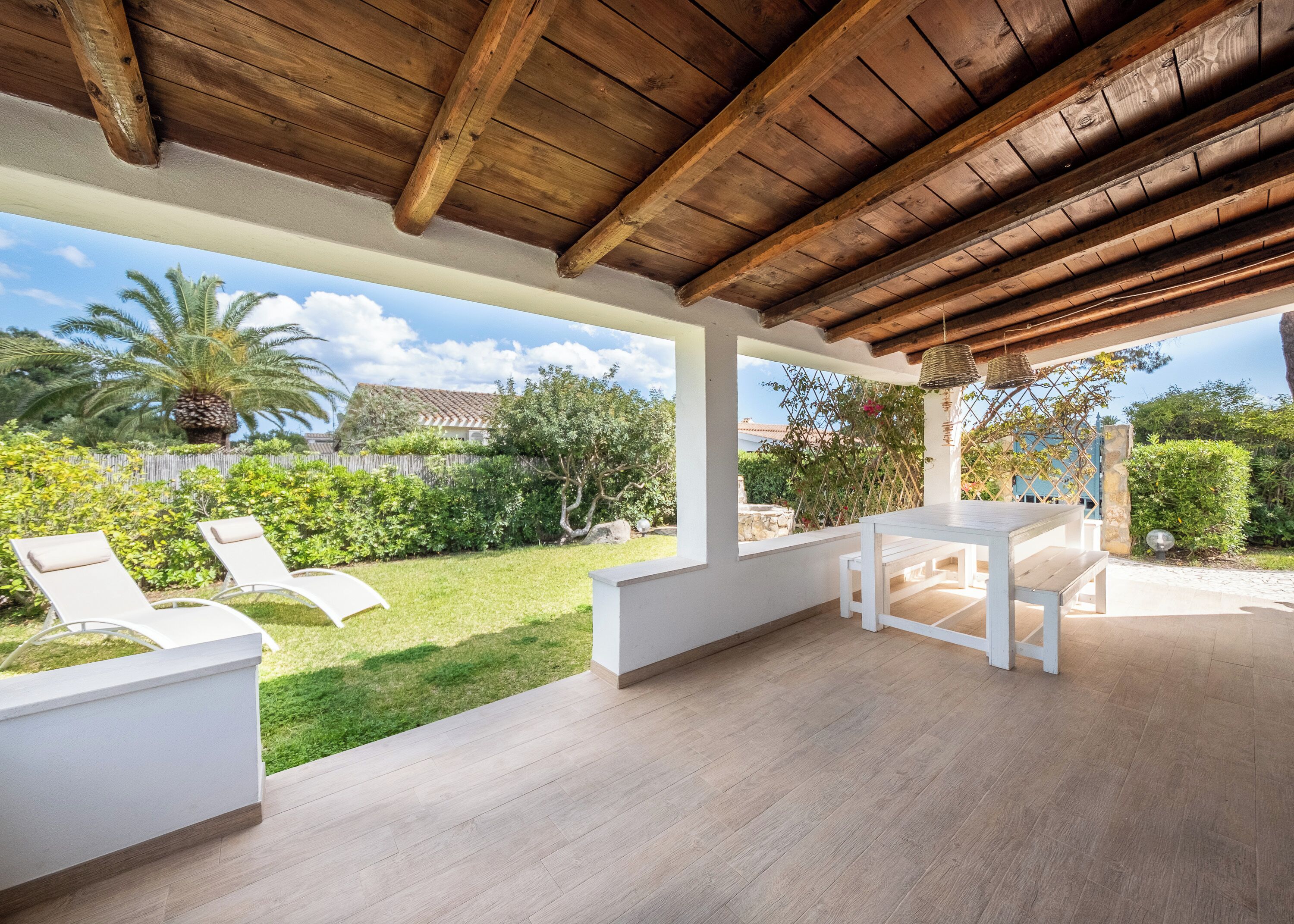 A covered patio with a wooden ceiling, white furniture, and a view of a garden with palm trees and a house in the background.