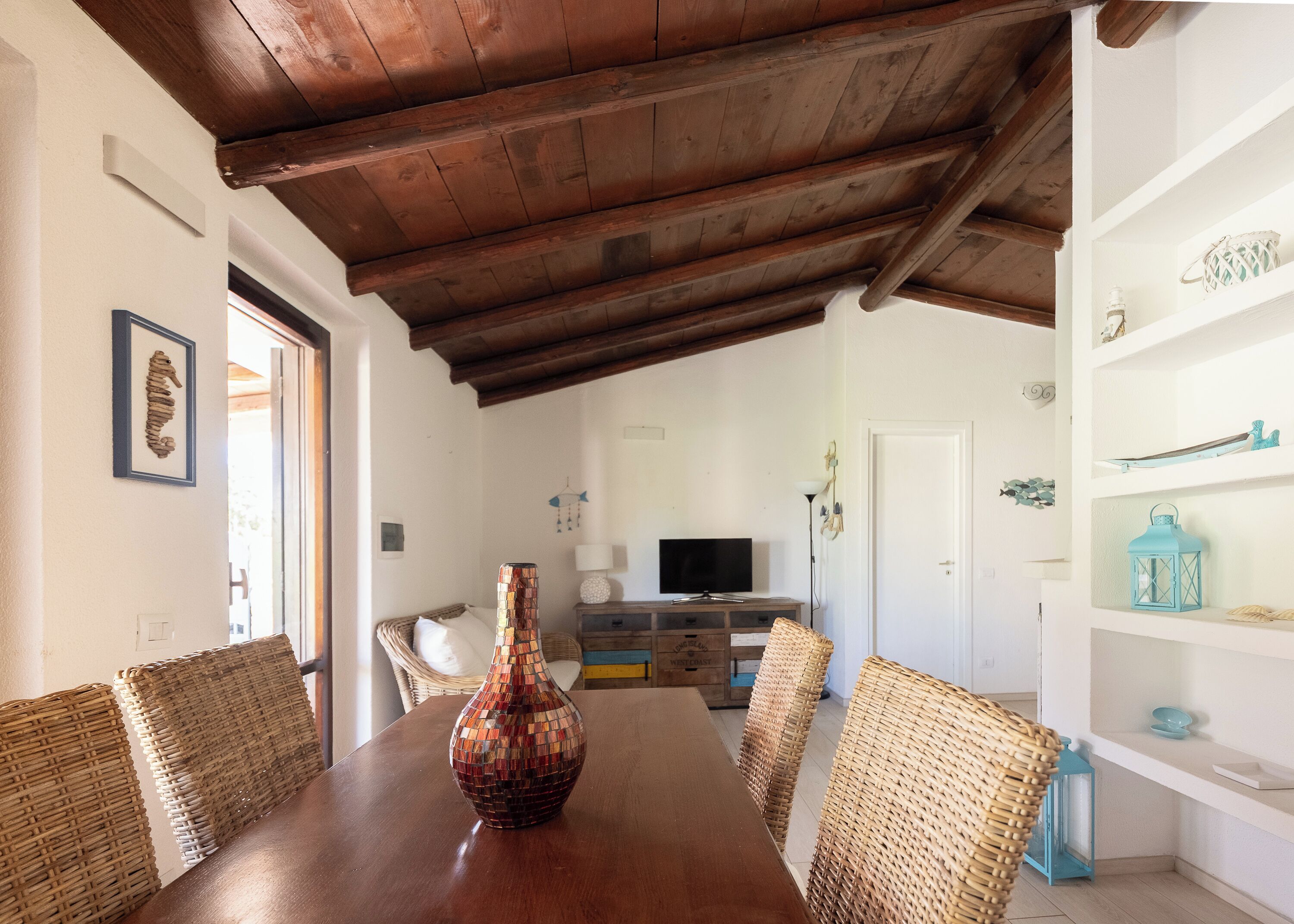 A dining area with a wooden table, wicker chairs, a decorative vase, and a television in the background.