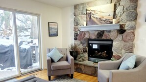 A living room with a stone fireplace, two armchairs, a sofa, and a view through a glass door.