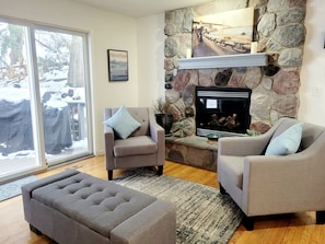 A living room with a stone fireplace, two armchairs, a sofa, and a view through a glass door.