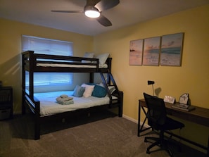 Uma cama de beliche com almofadas, uma escrivaninha com cadeira, um ventilador de teto e uma janela com persianas.