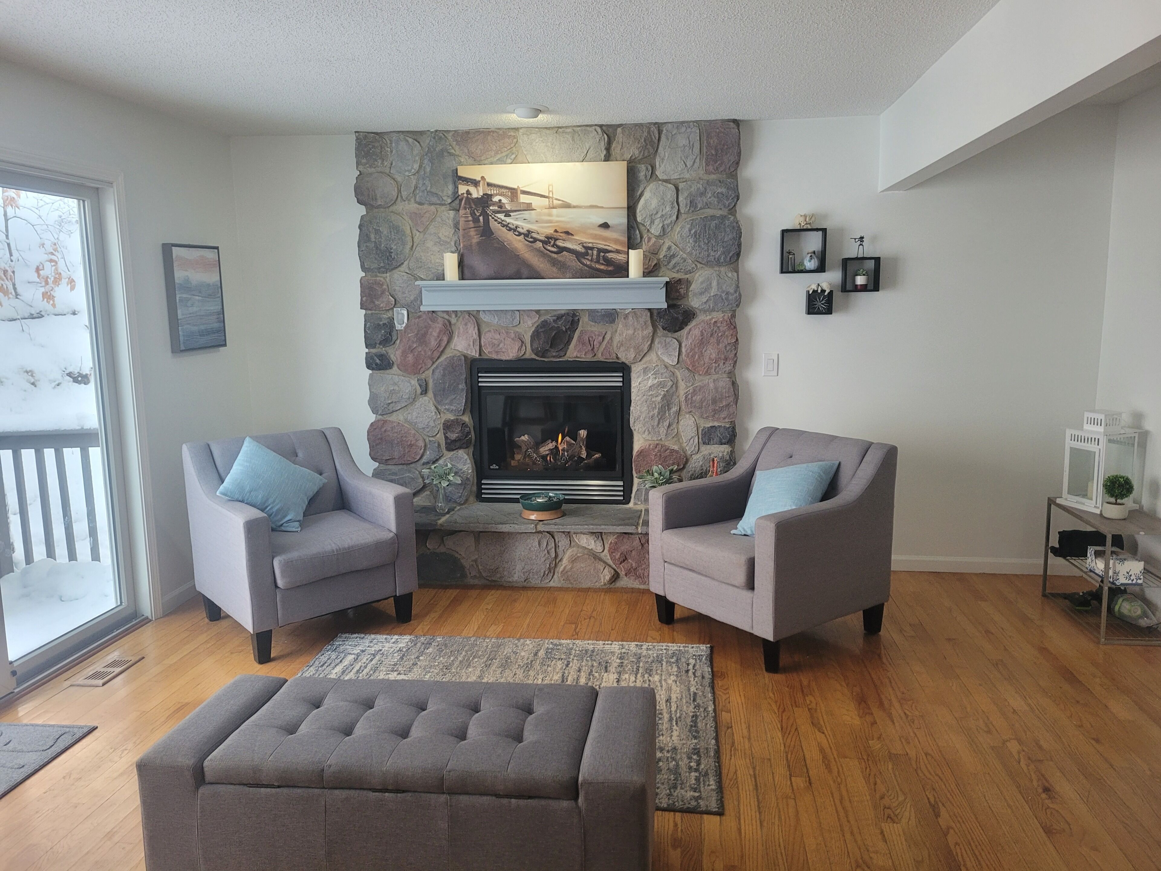 A living room with a stone fireplace, two armchairs, a bench, and a view of a bridge through the window.