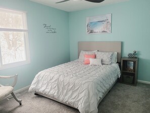 A bedroom with a bed, a nightstand, a chair, a window, and a wall with a quote.