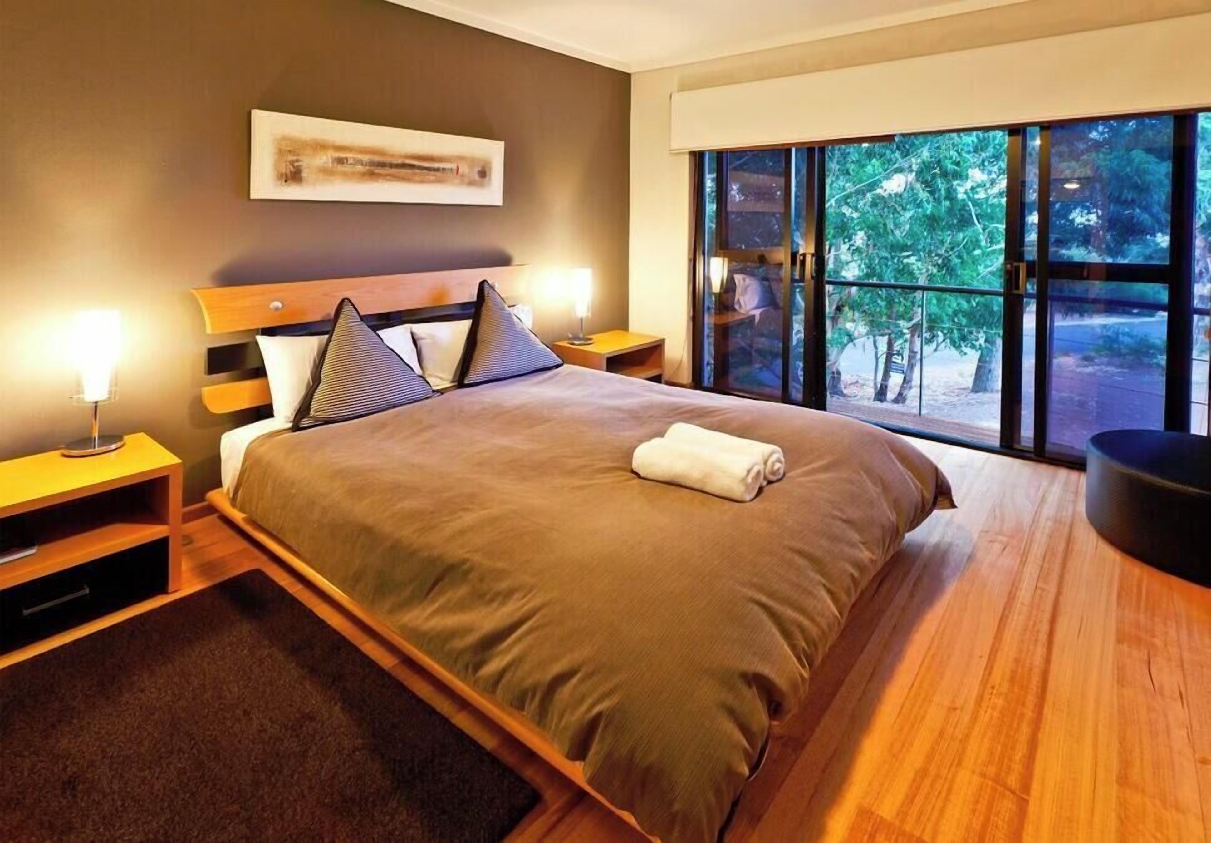 A modern bedroom with a large bed, two bedside tables, a nightstand, and a view of greenery through large windows.