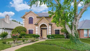 A two-story house with a brick facade, a central entrance with an arch, and a well-maintained front yard with a lawn and shrubs. - Private Pool Oasis- Spacious & Fully Furnished Dallas Home (Plano)