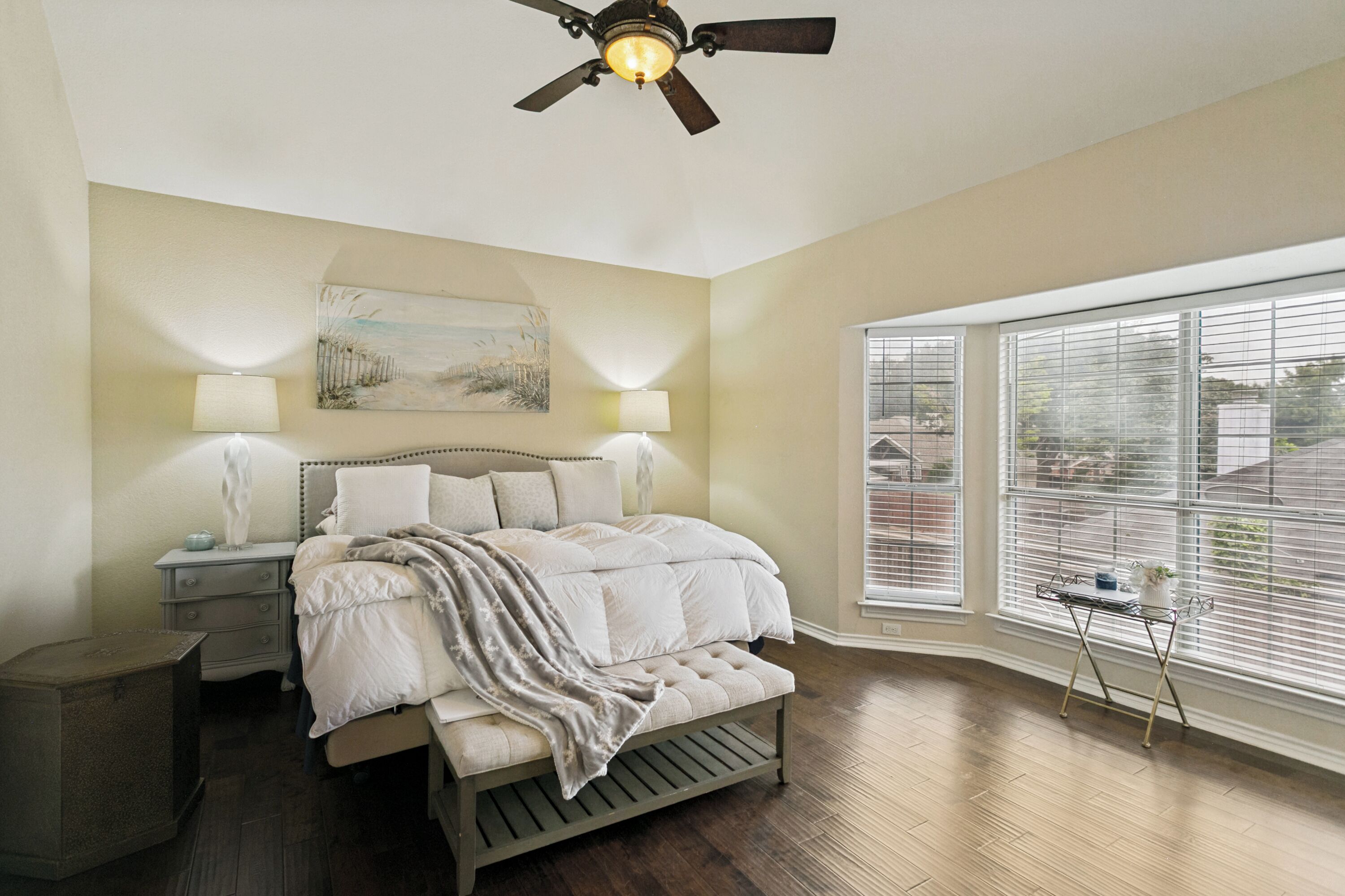 A bedroom with a bed, bedside tables, a ceiling fan, a window with blinds, and a painting on the wall.