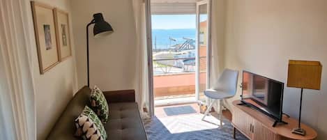 A hotel room with a sofa, a television, a lamp, and a view of the sea through an open door.