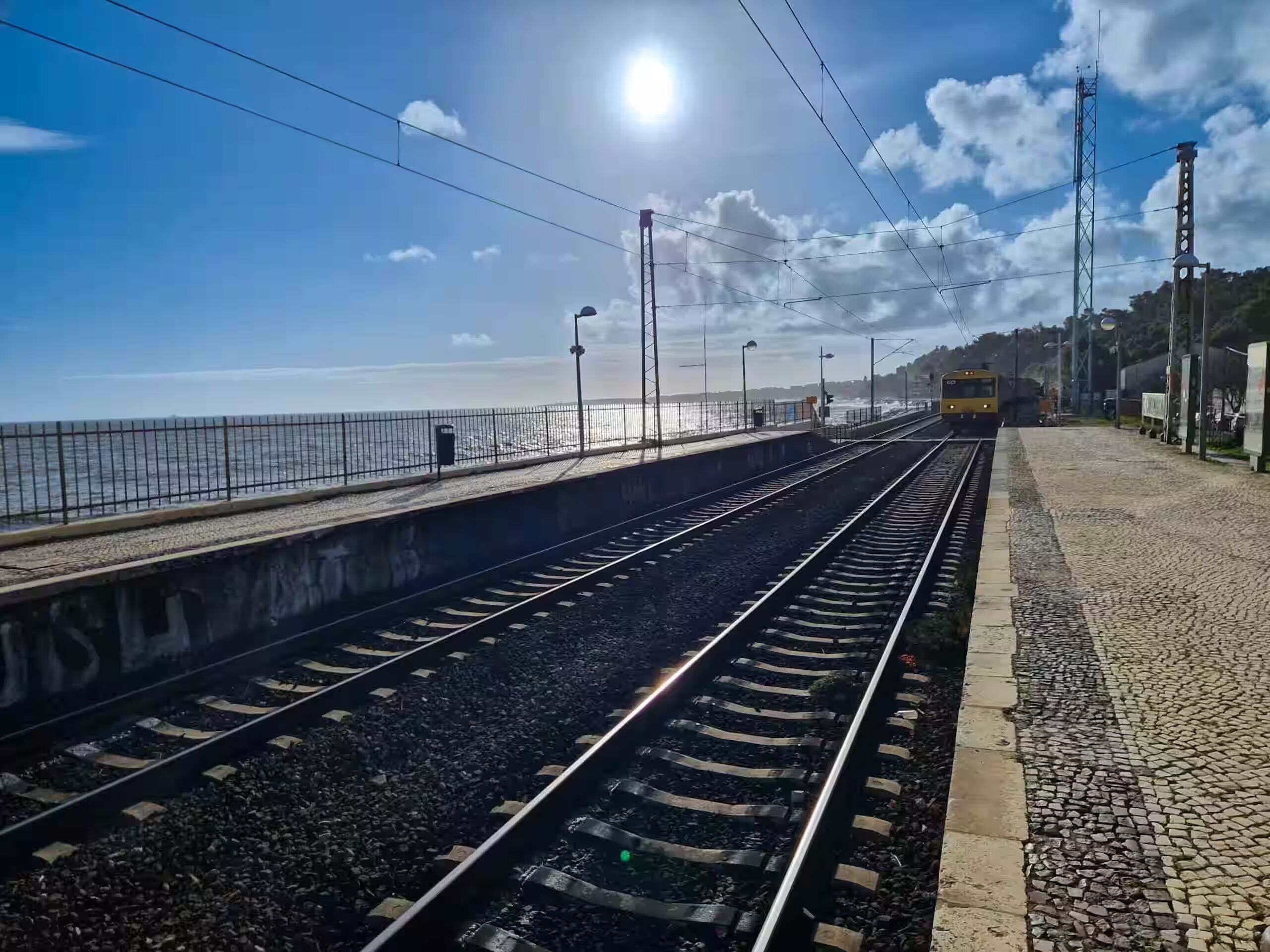 A train station platform with tracks, a fence, and a clear sky.