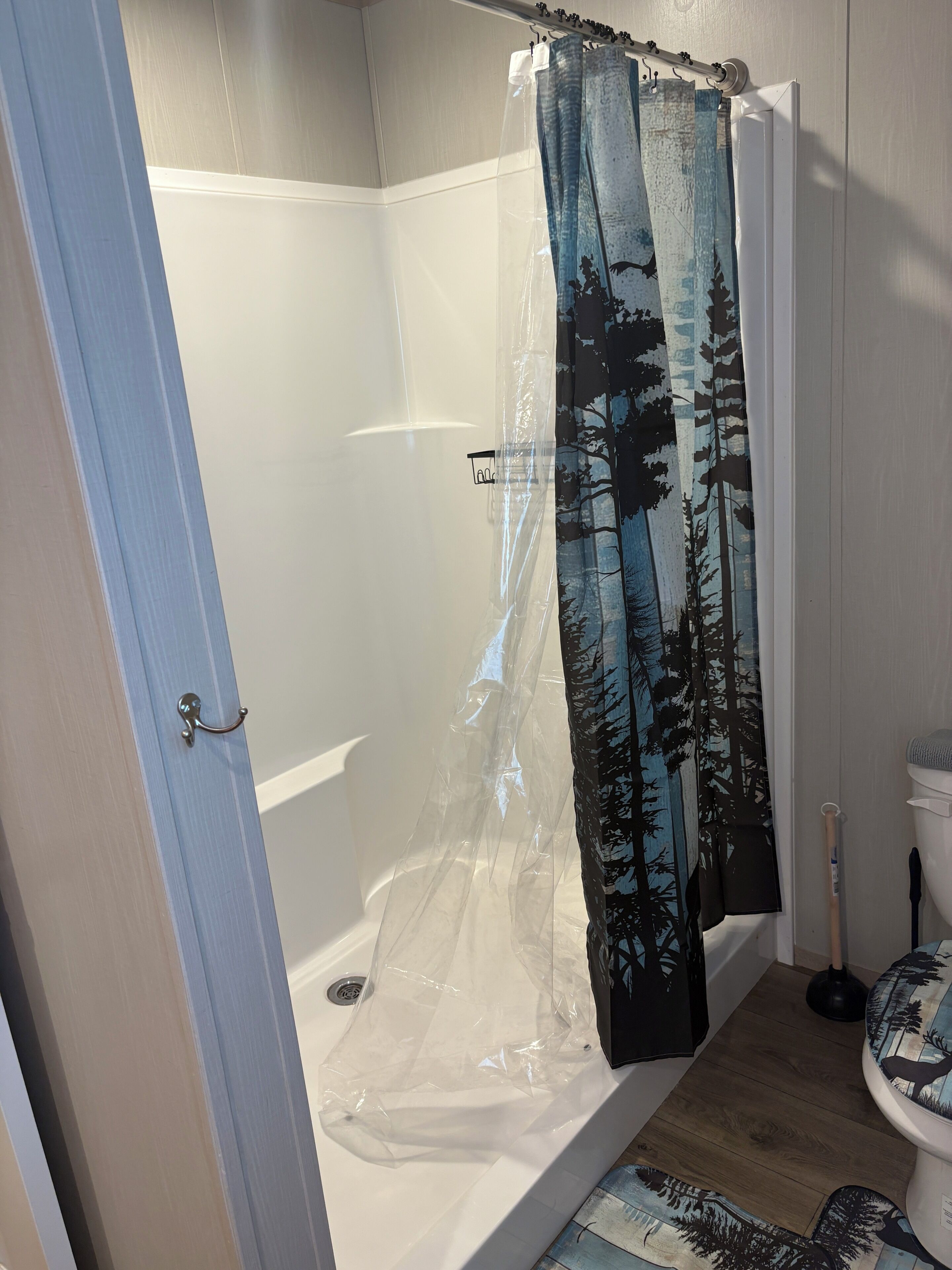 A bathroom with a shower curtain featuring a forest print, a white bathtub, and a wooden floor.