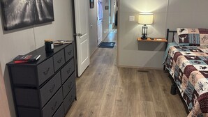 A bedroom with a bed, a bedside table with a lamp, a dresser with drawers, a door leading to a hallway, and a painting on the wall.