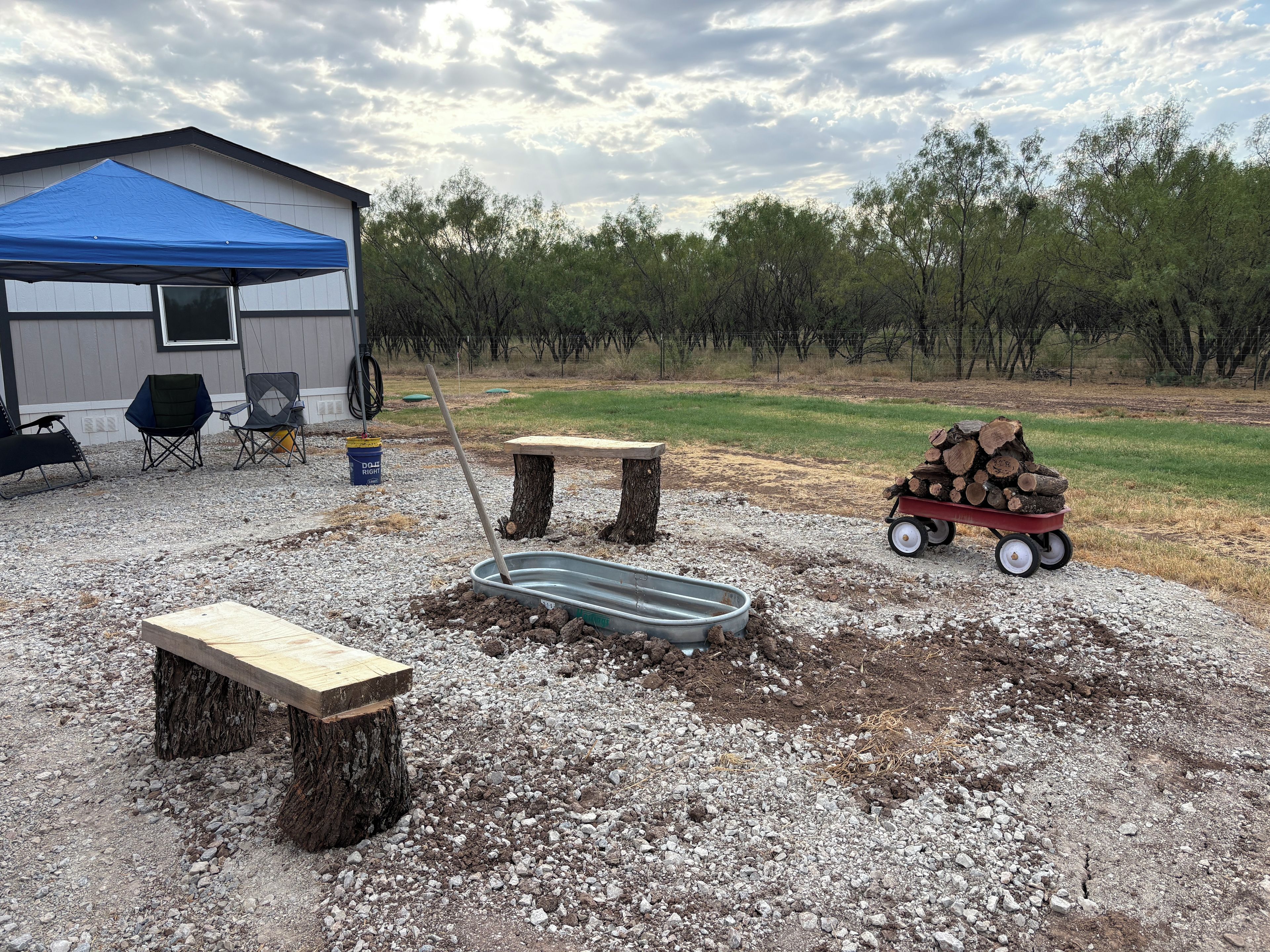 A covered outdoor area with a table, chairs, a bench, a wheelbarrow with logs, and a building with a blue roof.