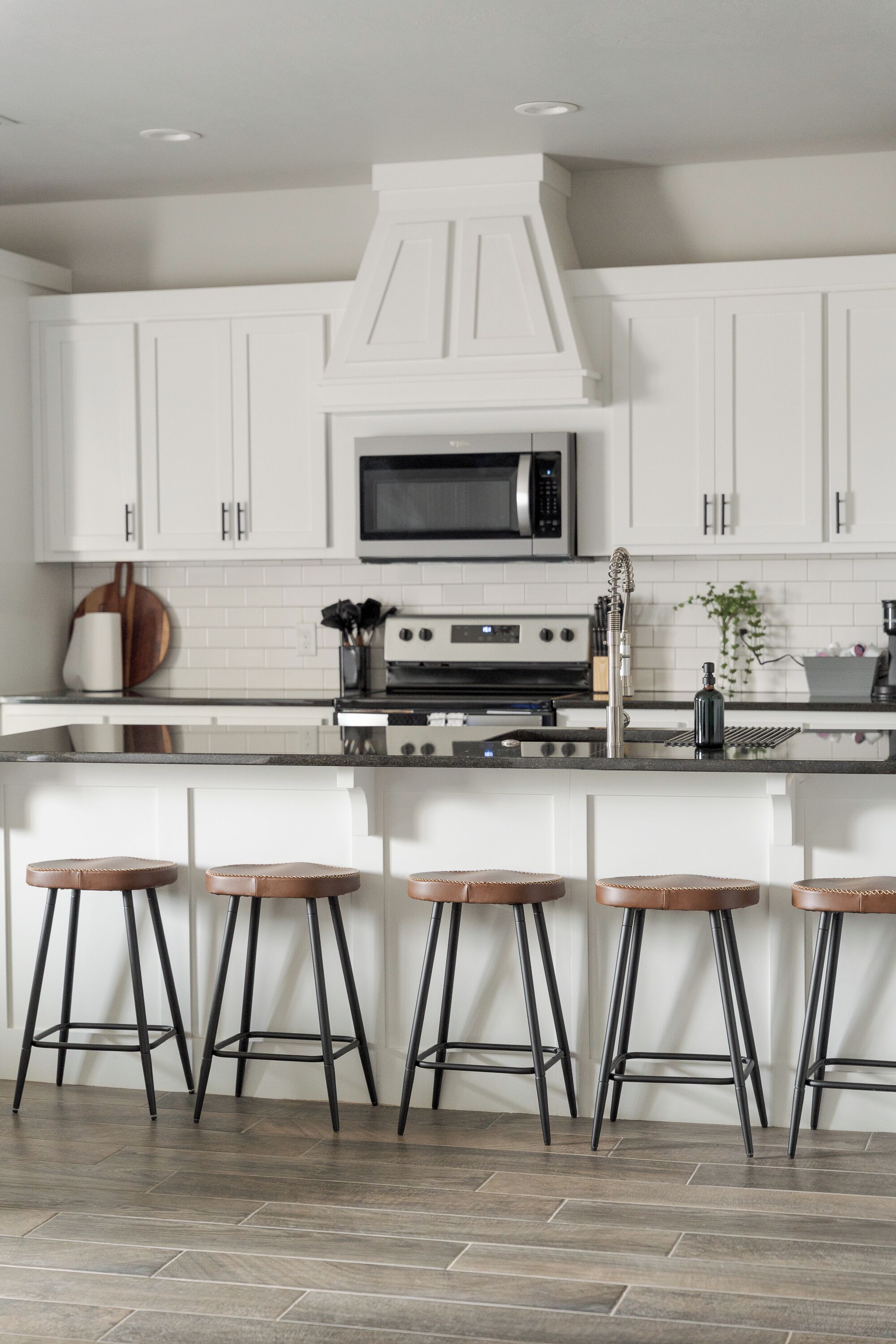 A kitchen with white cabinets, a black countertop, a stainless steel microwave, a gas stove, and four stools.