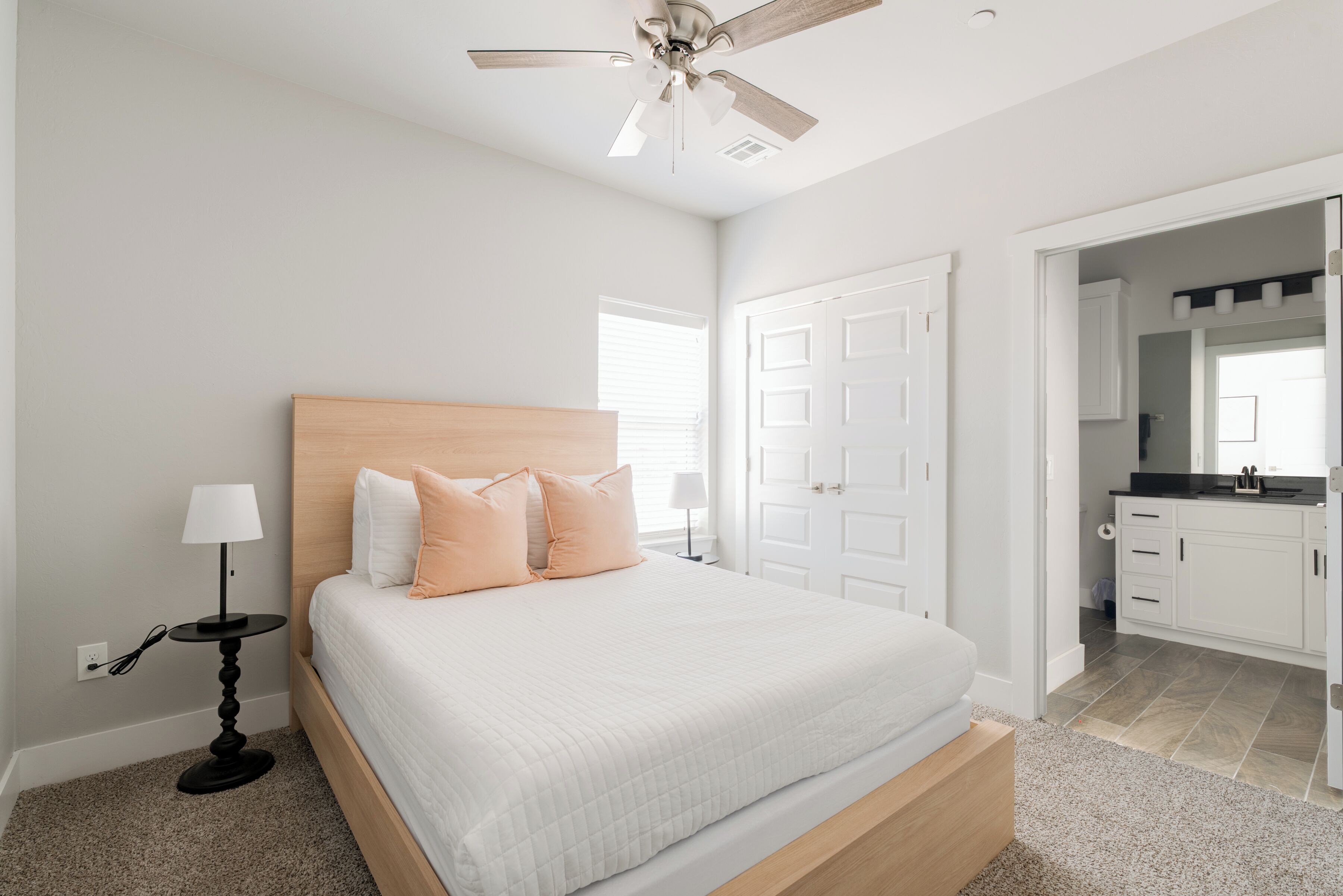 A bedroom with a bed, a nightstand, a lamp, a ceiling fan, and a view into the kitchen.
