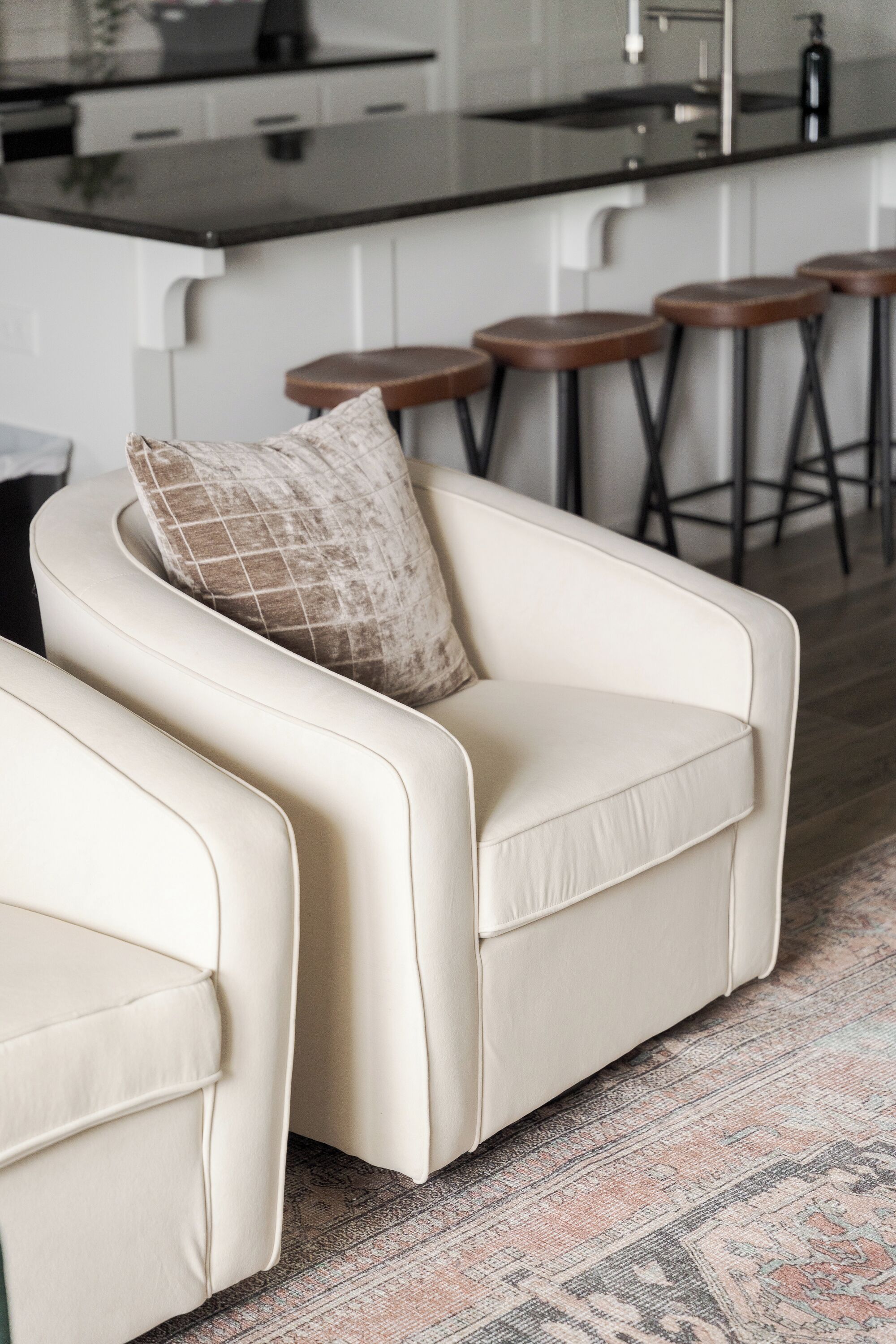 A modern living room with a sofa and a matching armchair, a patterned rug, and a kitchen area with bar stools in the background.