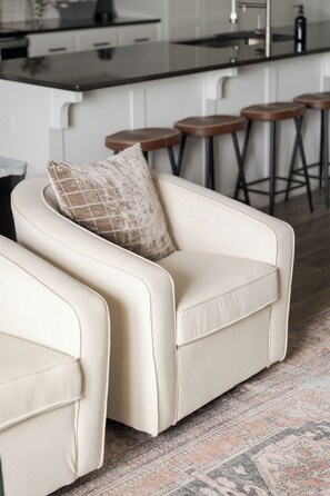 A modern living room with a sofa and a matching armchair, a patterned rug, and a kitchen area with bar stools in the background. - E201: 3BR near UCO | 2 King+ Private Bath Every BR (Edmond)