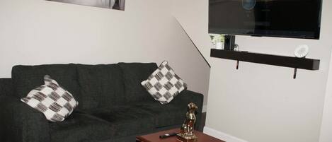 A living room with a grey sofa, two patterned cushions, a small wooden table with a remote control, a decorative dog statue, and a flat-screen TV mounted on the wall.
