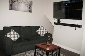 A living room with a grey sofa, two patterned cushions, a small wooden table with a remote control, a decorative dog statue, and a flat-screen TV mounted on the wall.