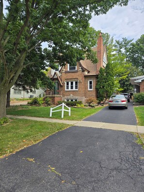 A residential street with a brick house, a driveway, and a car parked on the street. - American Tudor Revival, 8-beds, 4 bedrooms, 3 sofa beds, 3 full bath, all new. (Downers Grove)