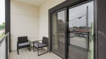 A balcony with two chairs and a small table, a sliding glass door, and a view of the street.