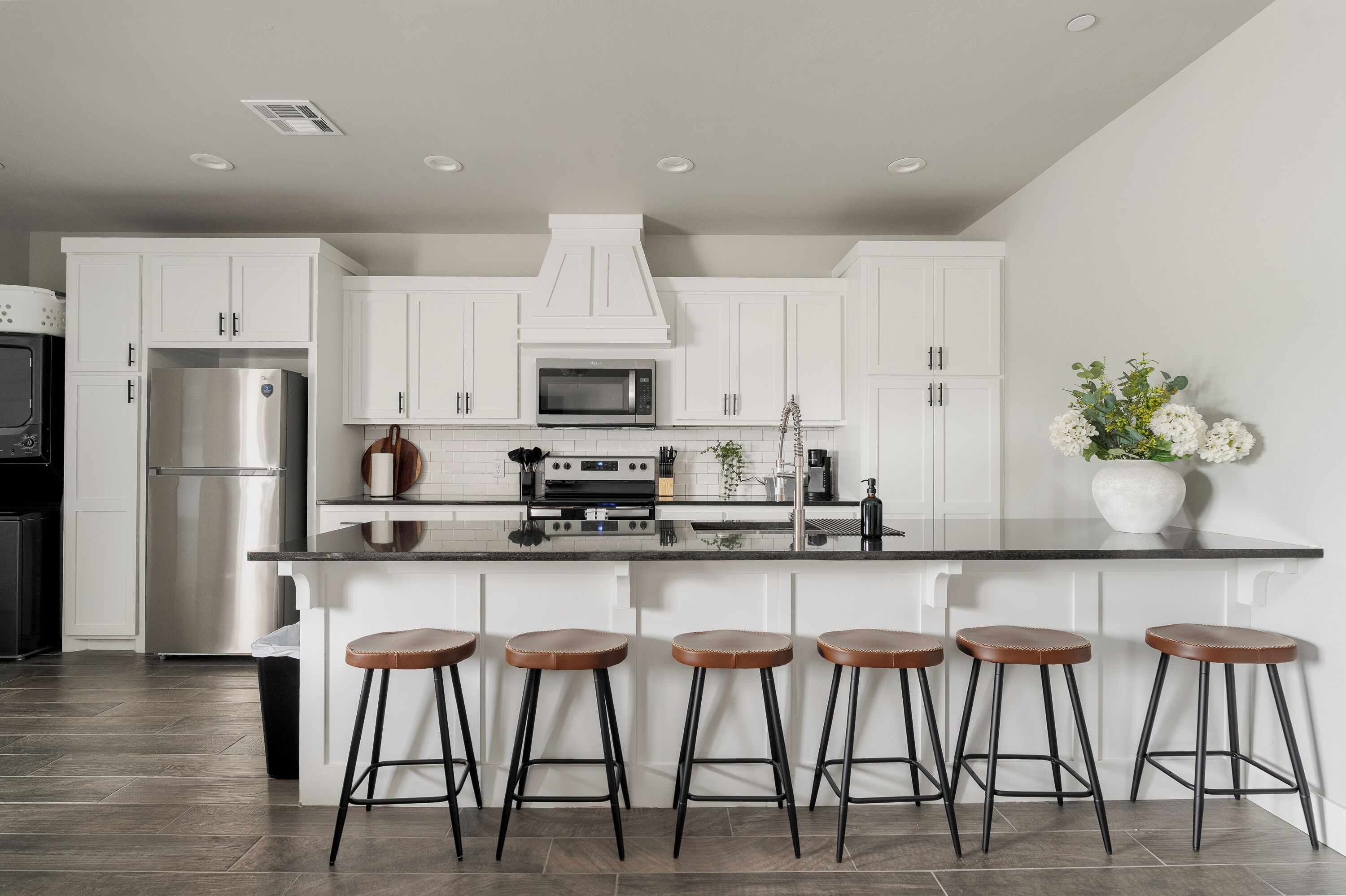 A modern kitchen with white cabinets, a stainless steel refrigerator, a microwave, and a built-in oven. There are four stools at the island and a vase with flowers on the counter.