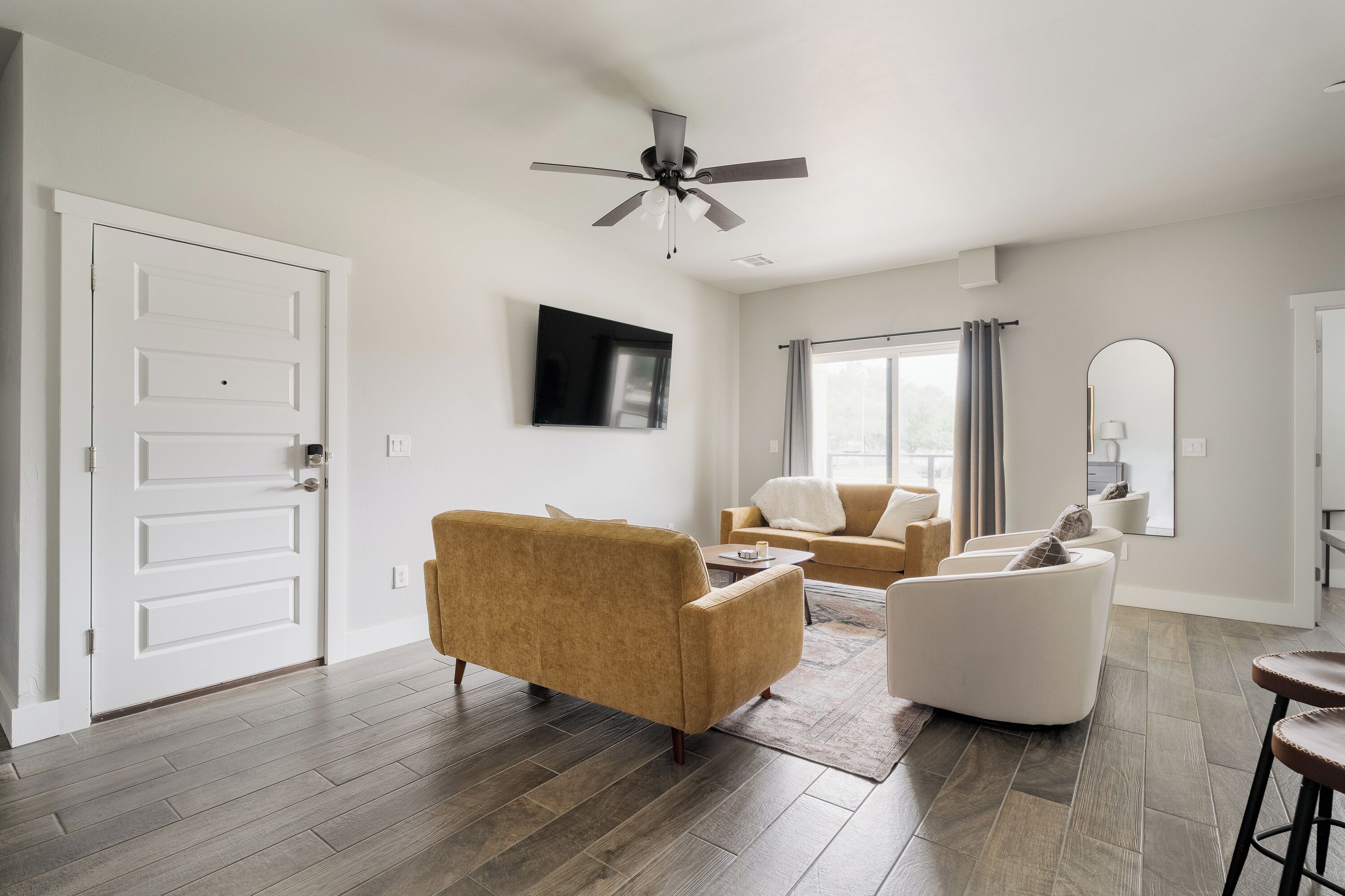 A modern living room with a ceiling fan, a flat-screen TV mounted on the wall, and a large window with curtains.