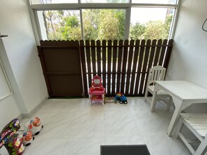 Una habitación con una mesa blanca, una silla blanca, un área de juegos pequeña con juguetes y vistas al exterior por una ventana.
