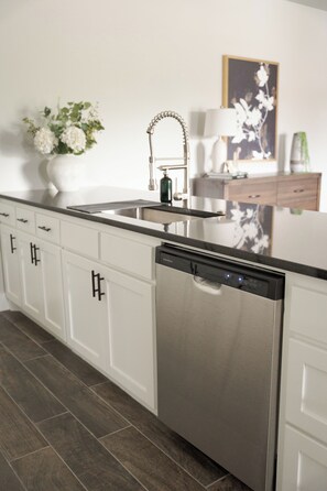 A modern kitchen with white cabinetry, a stainless steel dishwasher, and a black countertop. - E202: 3BR near UCO | 2 King+ Private Bath Every BR (Edmond)