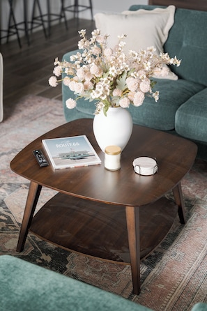 Una mesa redonda de madera con un jarrón blanco que contiene flores, un control remoto, una revista y un pequeño recipiente blanco.