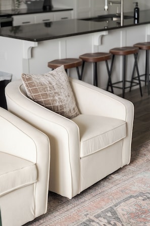A modern living room with a sofa and a matching armchair, a patterned rug, and a kitchen area with bar stools in the background.