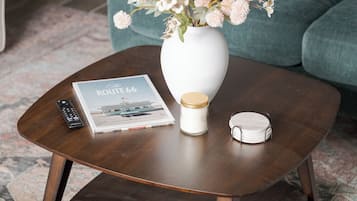A round wooden coffee table with a white vase containing flowers, a remote control, a magazine, and a small container.