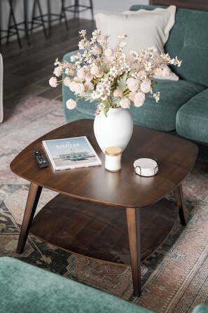 A round wooden coffee table with a white vase containing flowers, a remote control, a magazine, and a small container.