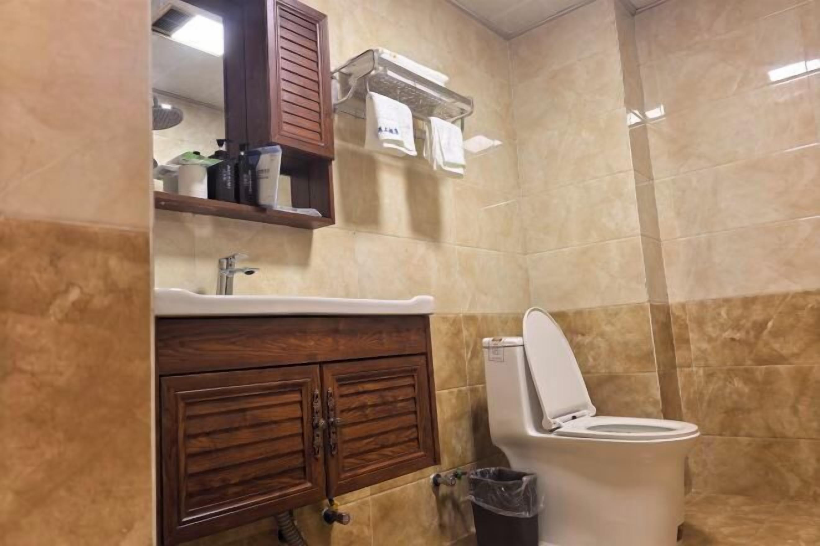 A bathroom with a toilet, sink, wooden cabinet, and a trash bin.