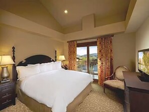 Habitación de hotel con una cama grande, un escritorio con televisor, una silla y vistas al exterior a través de una ventana con cortinas.