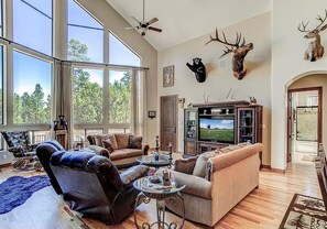 A spacious living room with a large television, comfortable seating, and a view of the outdoors through large windows.