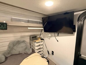 A room with a bed, a television mounted on the wall, a clock, and a small storage unit.