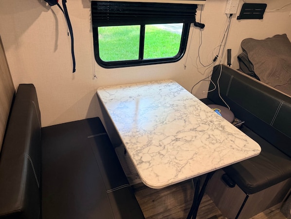 A compact RV interior with a marble-topped table, a window with a view of green grass, and a black leather bench.