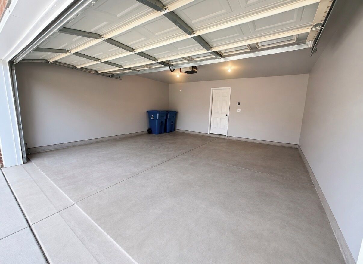 A garage with a concrete floor, a white door, and two blue trash bins.