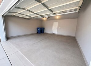 A garage with a concrete floor, a white door, and two blue trash bins.