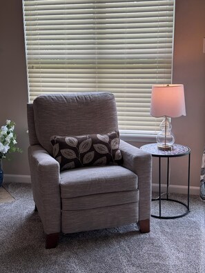 A grey armchair with a patterned pillow, a small table with a lamp, and a window with blinds.