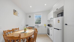 A modern kitchen with white cabinetry, a dining table with chairs, a window, and a clock on the wall. - Apple Court Retreat – 3 Bedroom – Access to Huon River (Huonville)